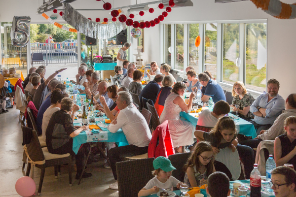 Une cinquantaine de personnes réparties autour de grandes tables en train de manger et discuter. La salle est décorée de guirlandes et de ballons. Au troisième plan, deux grosses enceintes de musique.
