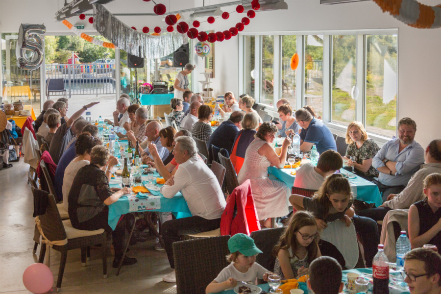 Une cinquantaine de personnes réparties autour de grandes tables en train de manger et discuter. La salle est décorée de guirlandes et de ballons. Au troisième plan, deux grosses enceintes de musique.